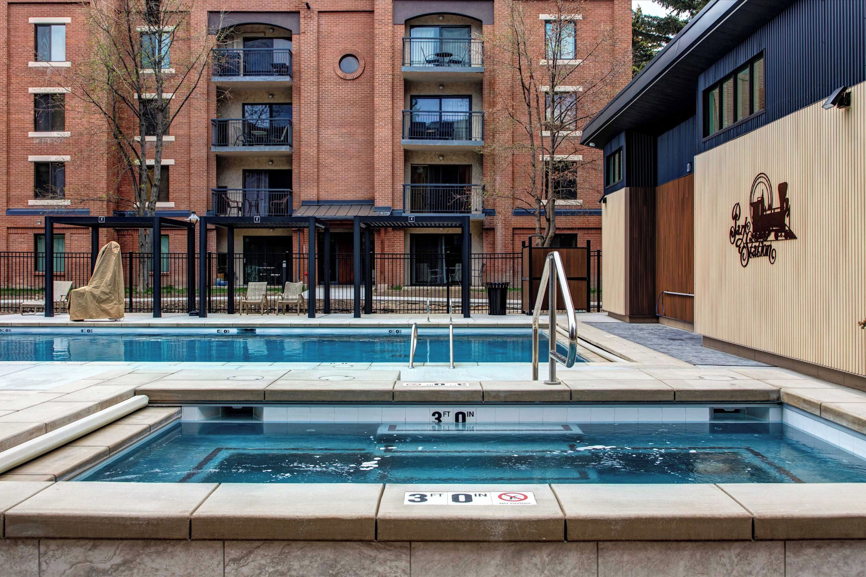 A swimming pool with a depth marker, surrounded by a brick building with balconies and a covered patio area.