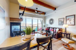 A spacious living area with a fireplace, dining table, and seating, featuring a large window and a ceiling fan. - Dog-friendly Newpark townhome with  private hot tub, washer & dryer, & central AC (Park City)