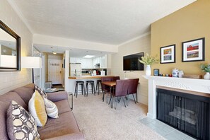 Ein modernes Wohnzimmer mit Sofa, Couchtisch und Kamin. Im Hintergrund ist eine Küche mit einer Bar zu sehen.