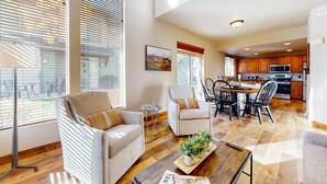Amplia sala de estar con sofá, sillón y mesa de comedor, una cocina con armarios de madera y ventanas grandes con persianas.