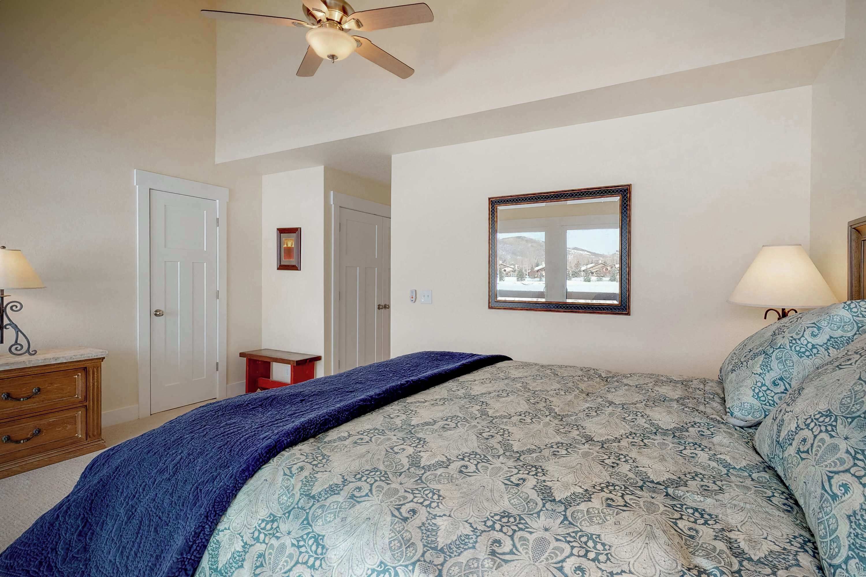A bedroom with a bed, a ceiling fan, a window with a view, and a door.