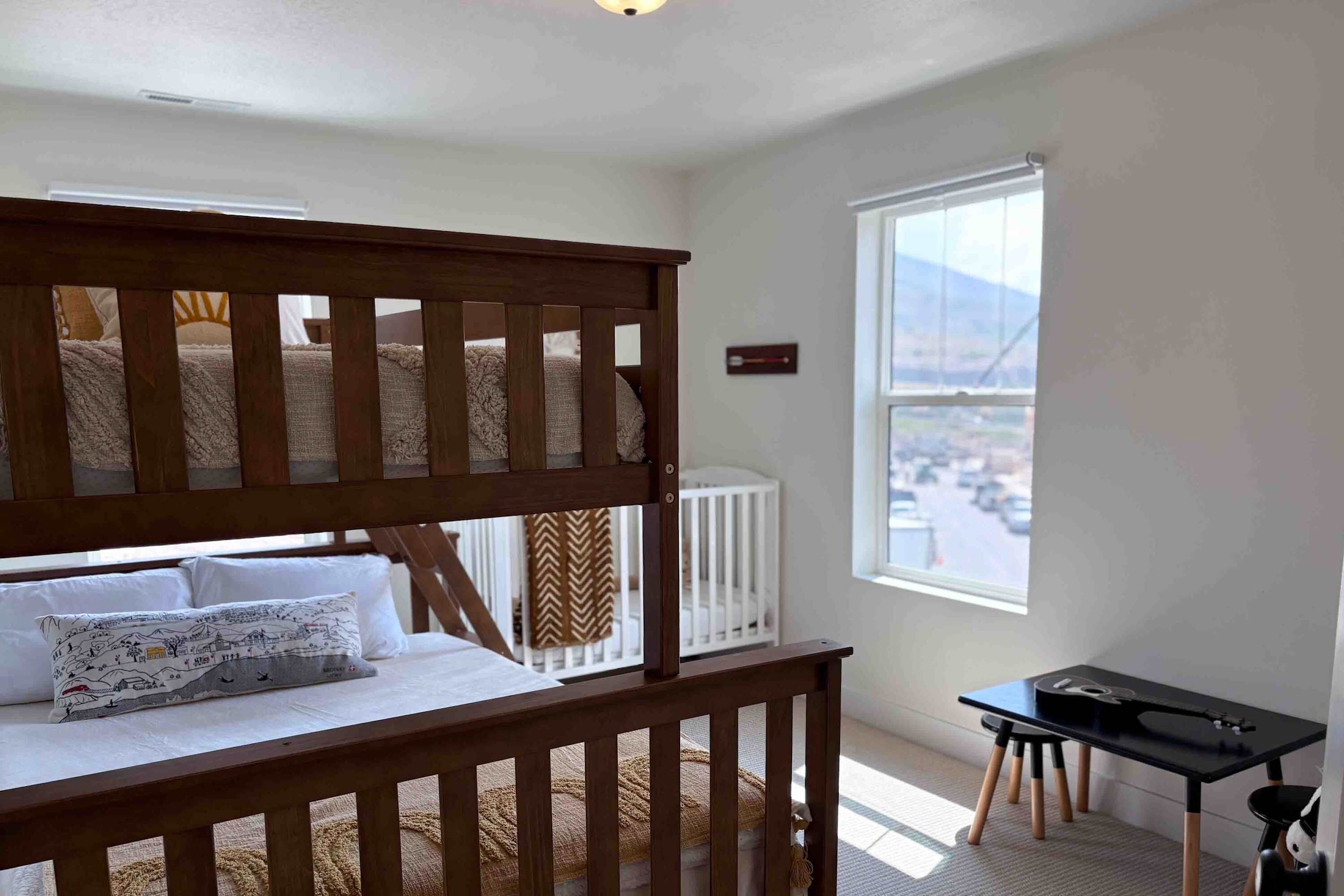 Quarto com cama de solteiro, berço, mesa pequena e janela com vista.