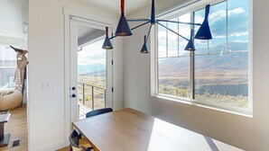 Ruokailutila, jossa on puinen pöytä ja tuolit, suuri ikkuna, josta avautuu näkymä maisemaan, sekä kattovalaisimet.