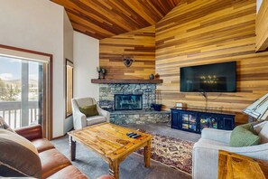 A living room with a stone fireplace, wooden ceiling, and a flat-screen TV mounted on the wall. - Lodge-Inspired Lakeside Townhouse with Fireplace - Short Walk to Lifts (Park City)