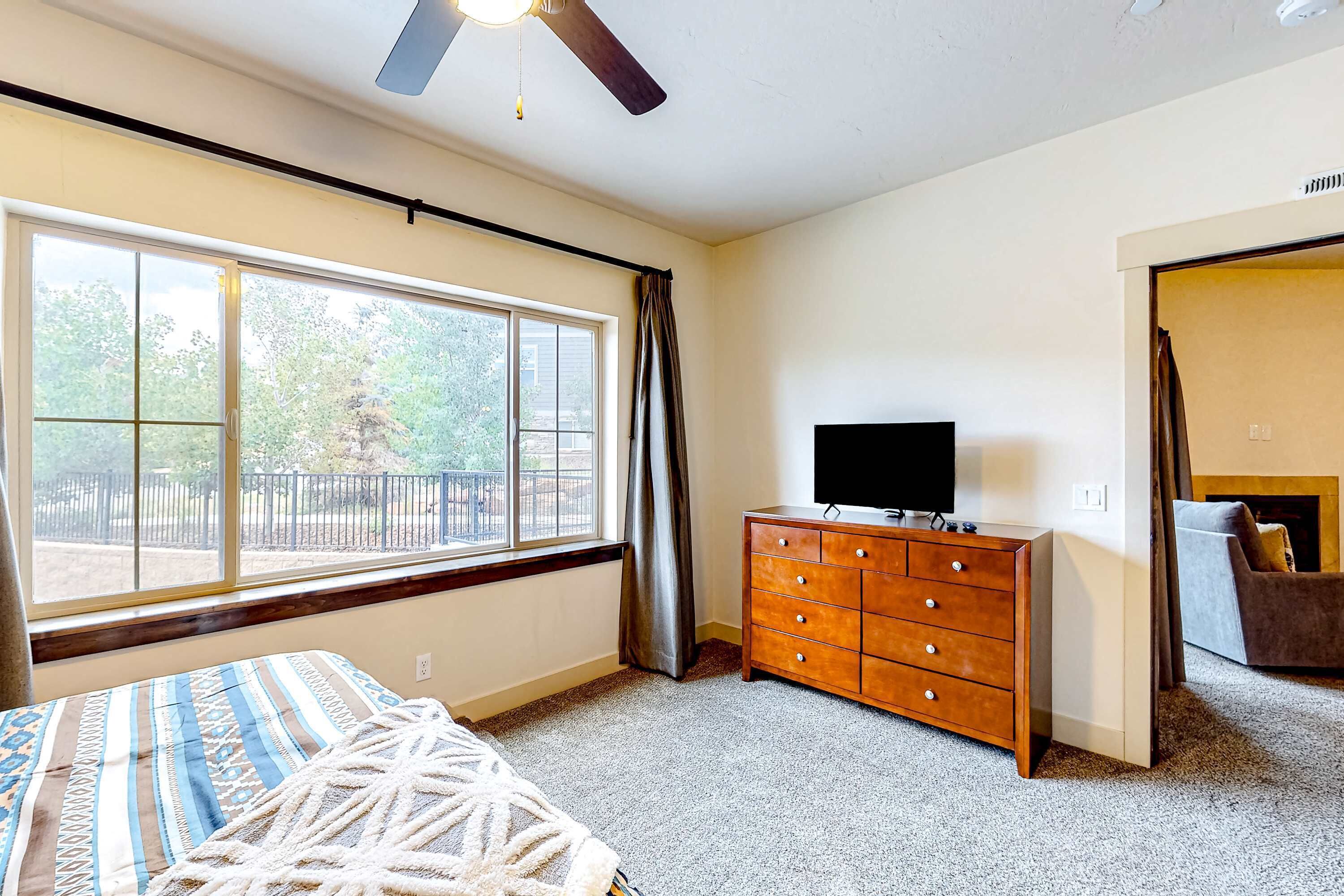 A bedroom with a bed, a dresser, a television, and a window with a view of trees.
