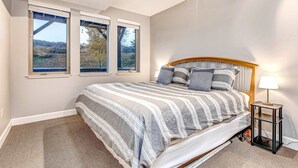 Ein Schlafzimmer mit einem großen Bett, drei Fenstern, einem Nachttisch mit Lampe und Blick ins Freie.