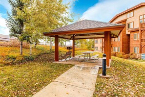 Un espace extérieur abrité, doté d’une table de pique-nique et de bancs, situé à côté d’un bâtiment à la façade en bois.