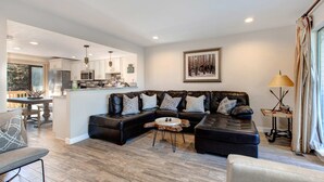 A modern living room with a sectional sofa, a coffee table, and a dining area with a kitchen in the background. - Three-story townhome with private hot tub, gas fireplace, decks (Park City)