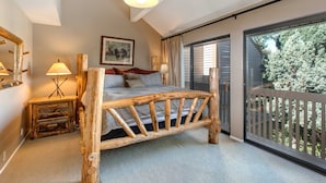 A bedroom with a wooden bed, a nightstand, a lamp, a painting, and a balcony with a view of trees. - Three-story townhome with private hot tub, gas fireplace, decks (Park City)