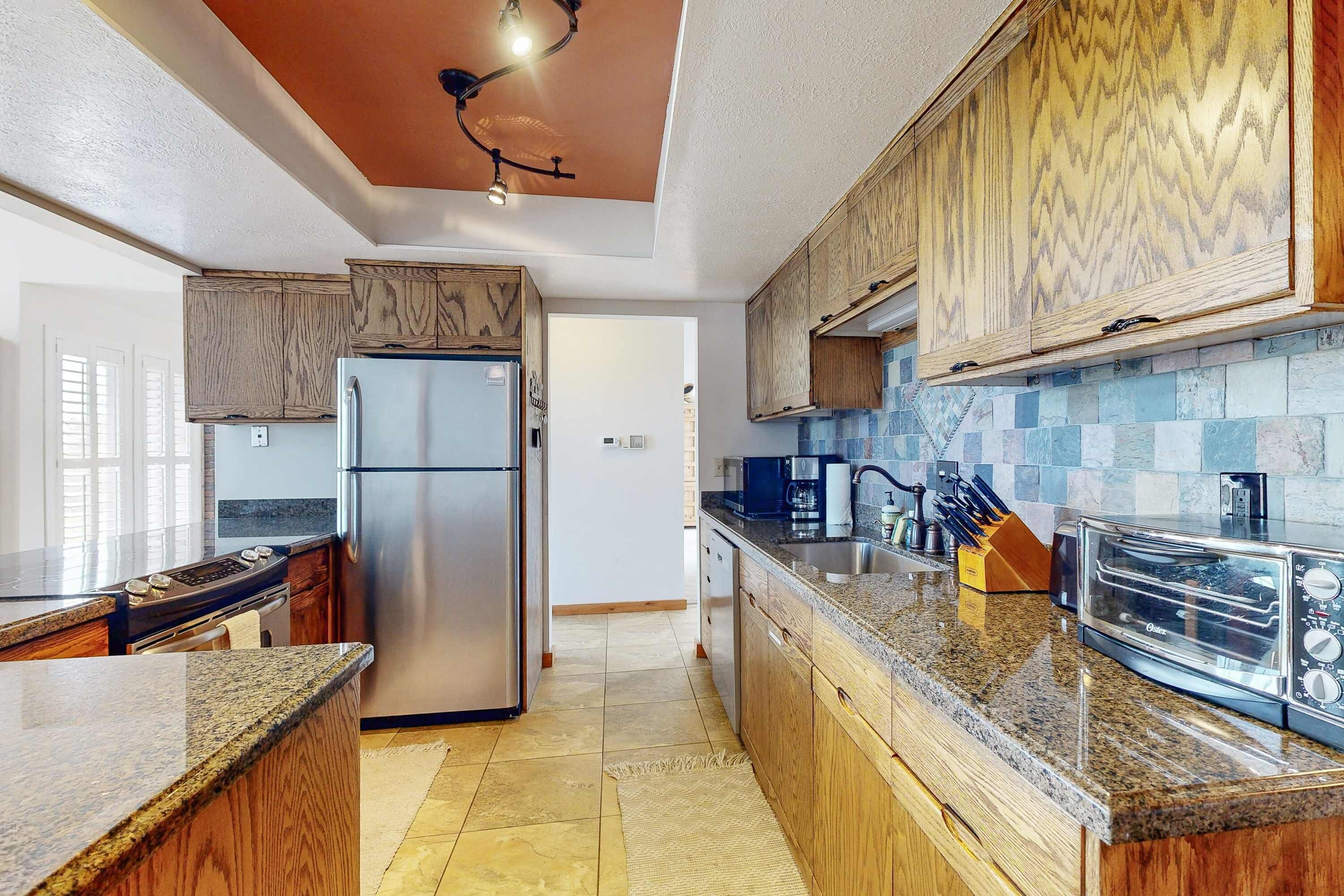 A kitchen with wooden cabinets, a stainless steel refrigerator, granite countertops, and a tiled backsplash.