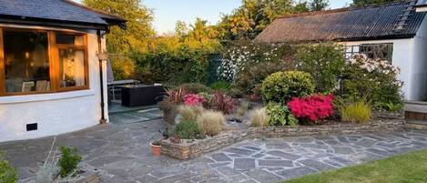A stone patio with a grassy area, a white building with a window, and a garden with various plants.