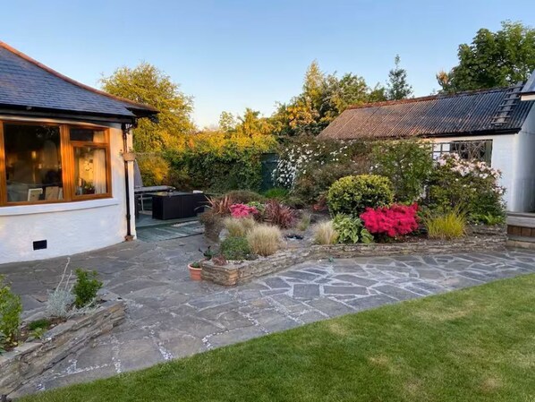 A stone patio with a grassy area, a white building with a window, and a garden with various plants.