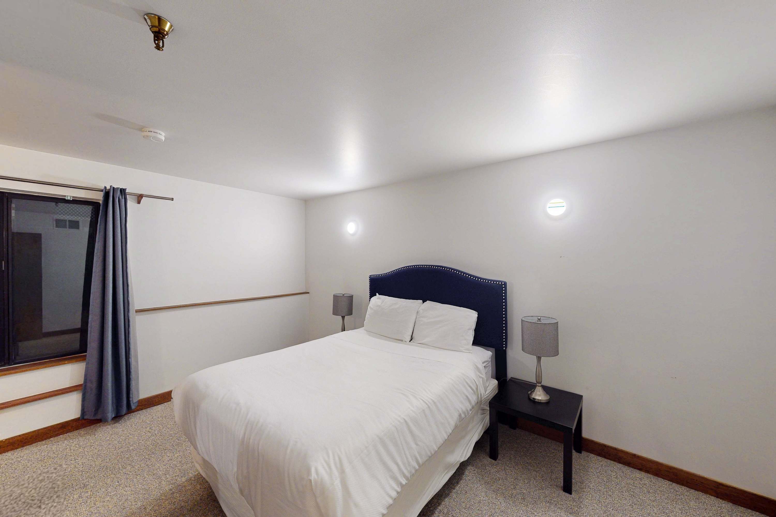 Una camera da letto con un letto, un comodino, una lampada, una finestra con tende e una luce a soffitto.