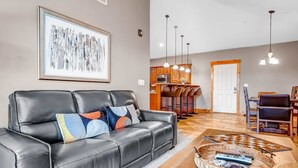A living room with a black leather sofa, a coffee table with a chessboard, a framed abstract painting, and a kitchen in the background. - Ski view condo with fast WiFi, private grill, easy ski access, pool, & hot tub (Park City)