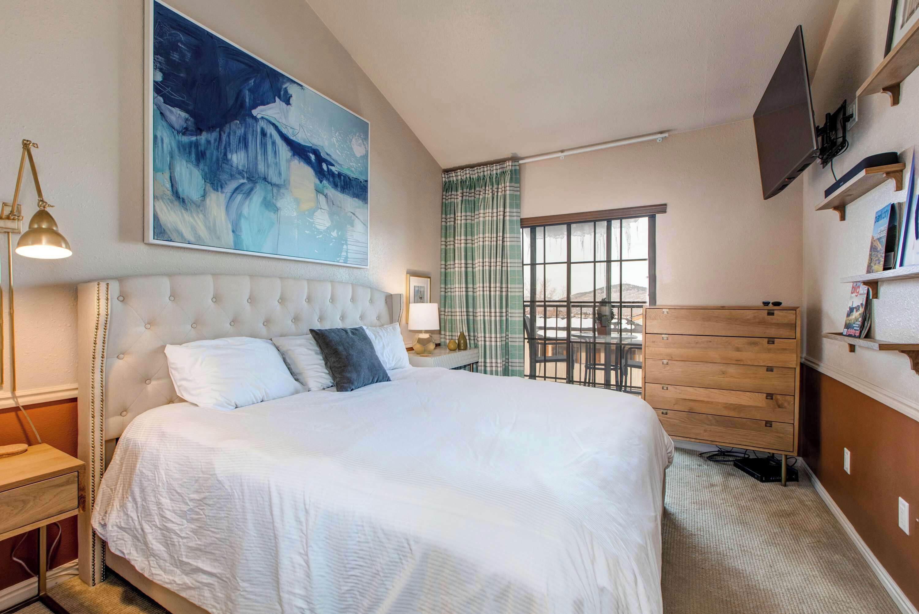 Une chambre à coucher avec un grand lit, une table de chevet, une lampe, une commode, un téléviseur et une fenêtre donnant sur un beau paysage.