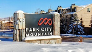 Park City Mountain -vuoristokeskukseen johtava opaste, jossa on logo. Opasteen ympärillä on lunta ja rakennuksia.