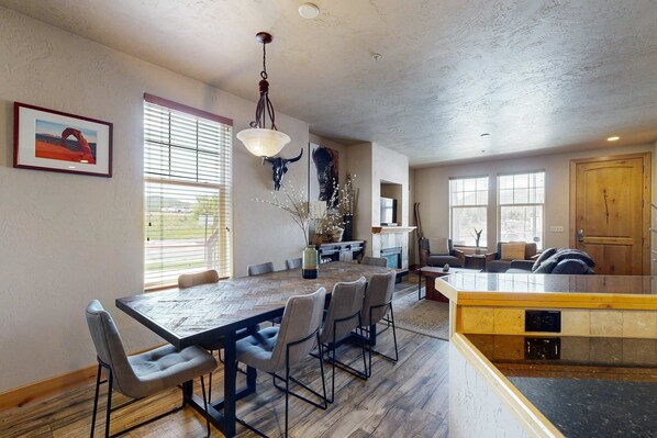 A spacious dining area with a large wooden table and chairs, a hanging light fixture, and a view of the kitchen area.