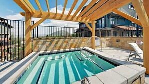 Uma área de piscina com pérgola de madeira, paredes de pedra e vista para uma casa.