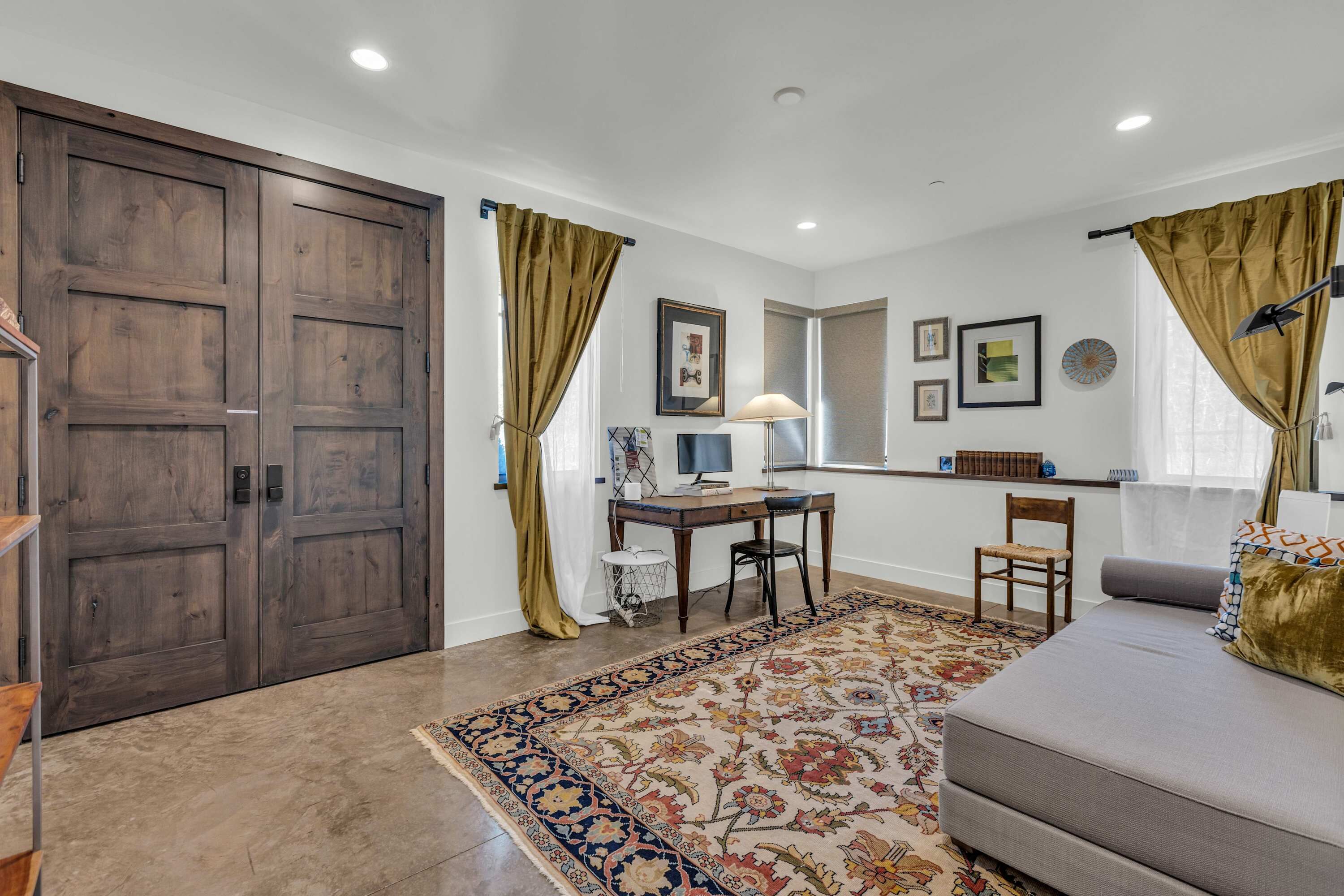 A room with a large wooden door, a desk with a computer, a sofa, and a patterned rug.