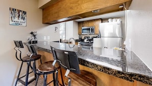 Uma cozinha moderna com bancada de granito, armários de madeira, geladeira de aço inoxidável e micro-ondas. Há dois banquetas de bar e uma placa indicando 'Park City, Utah'.