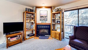 Una sala de estar con chimenea, estanterías de madera, un televisor y un ventanal con vistas a árboles.