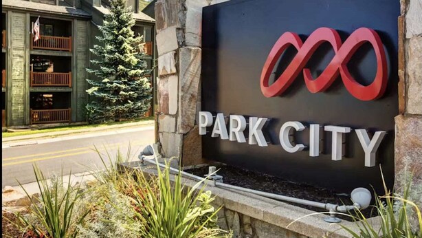 Señalización de 'PARK CITY' con un logotipo, frente a una pared de piedra y un edificio al fondo.