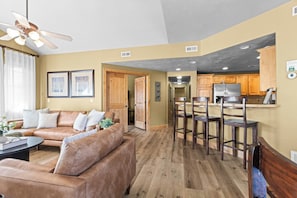 A living room with a brown leather sofa, a coffee table, and a ceiling fan. There is a kitchen area with wooden cabinets and a refrigerator. - Spacious Redstone Home with Fireplace, Patio & Walk to Dining (Park City)