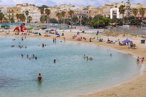 Une plage où l’on voit des gens nager et bronzer, une tour de surveillance des sauveteurs, et des bâtiments en arrière-plan.
