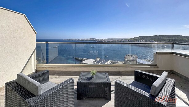 Un balcone con mobili in vimini, ringhiera in vetro e vista sul mare e colline lontane.