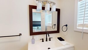 Ein Badezimmer mit einem weißen Waschbecken, einem Spiegel mit braunem Rahmen, einem Handtuchhalter und einem Fenster mit Jalousien.