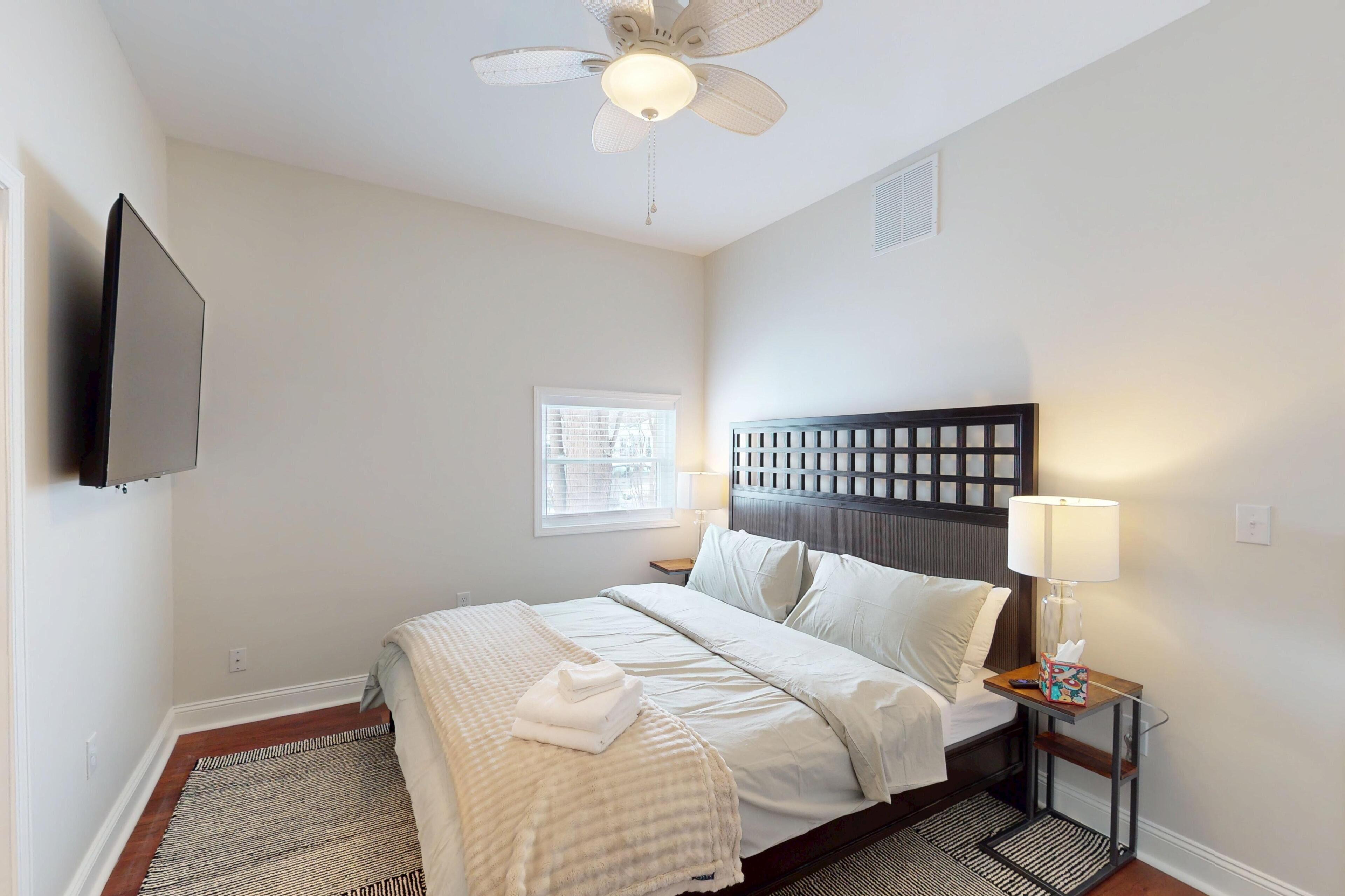 A hotel room with a bed, a bedside table with a lamp, a ceiling fan, and a TV mounted on the wall.