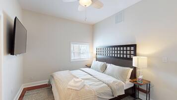 A hotel room with a bed, a bedside table with a lamp, a ceiling fan, and a TV mounted on the wall.