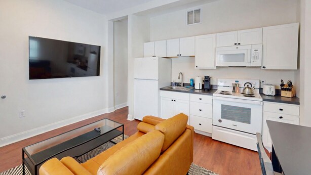 Eine moderne Wohnung mit Wohnzimmer, Sofa, Flachbildfernseher, Küche mit weißen Schränken, Mikrowelle und Kaffeemaschine.
