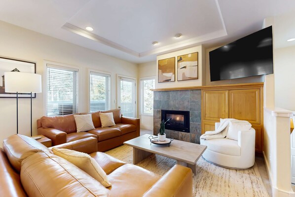 A living room with leather sofas, a fireplace, a flat-screen TV, and a large window. - Conveniently located townhome with kitchen, kitchenette, balcony & fireplace (Bend)