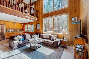A spacious living room with a large sectional sofa, a coffee table, and a fireplace. There is a kitchen area with wooden cabinets and a refrigerator. Large windows provide views of the surrounding forest.