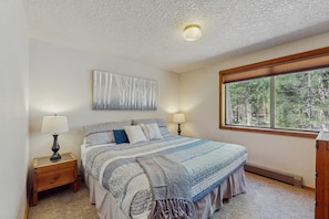 A bedroom with a large bed, two bedside tables with lamps, a ceiling light, a window with a view of trees, and a wall-mounted abstract painting.