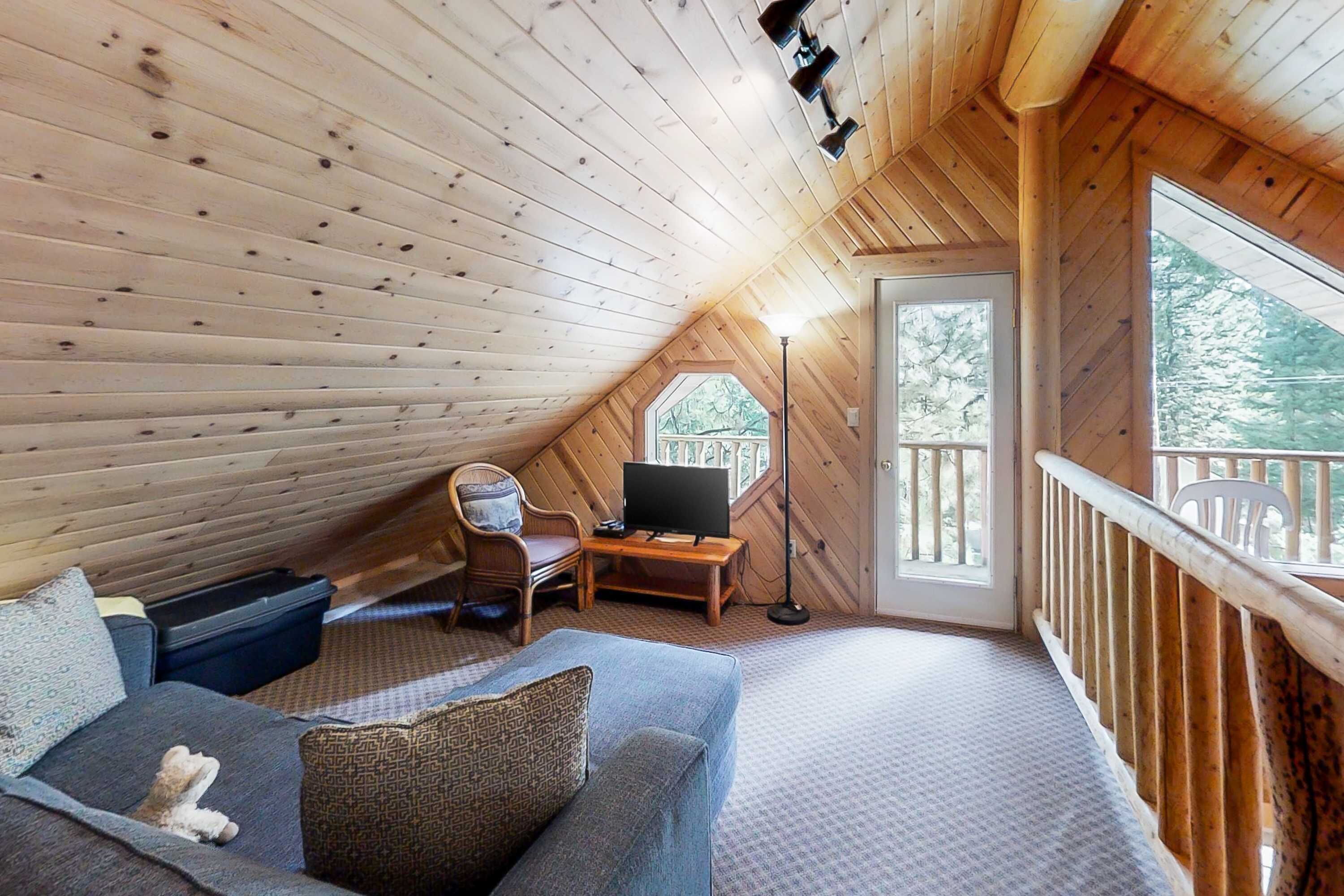 A cozy attic bedroom with a wooden ceiling, a sofa, a television, and a window overlooking greenery.
