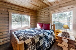 Un dormitorio de cabaña de madera con una cama, almohadas, una mesita de noche, una lámpara y una ventana.