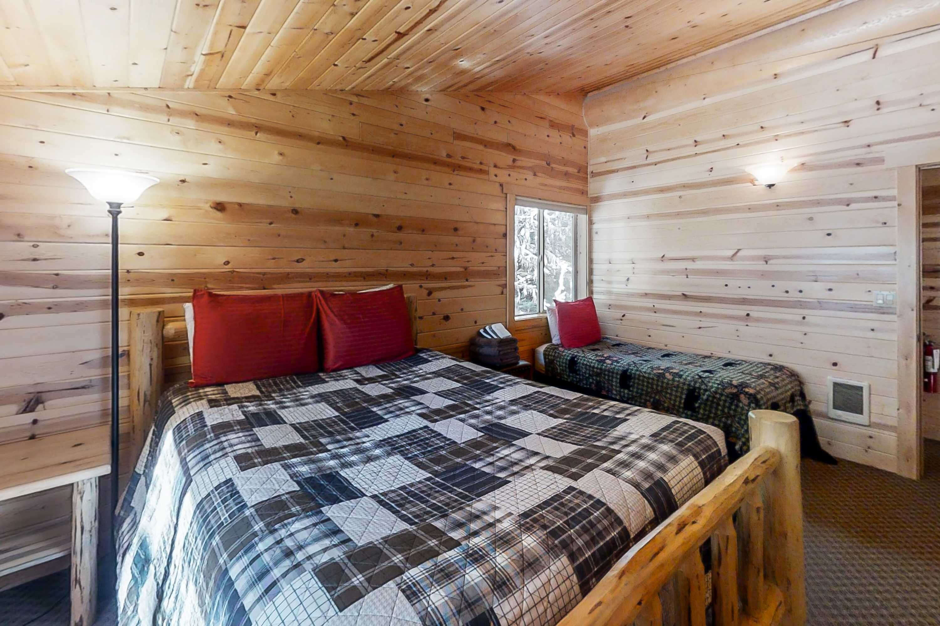 A wooden cabin room with a bed, two smaller beds, a window, and a lamp.