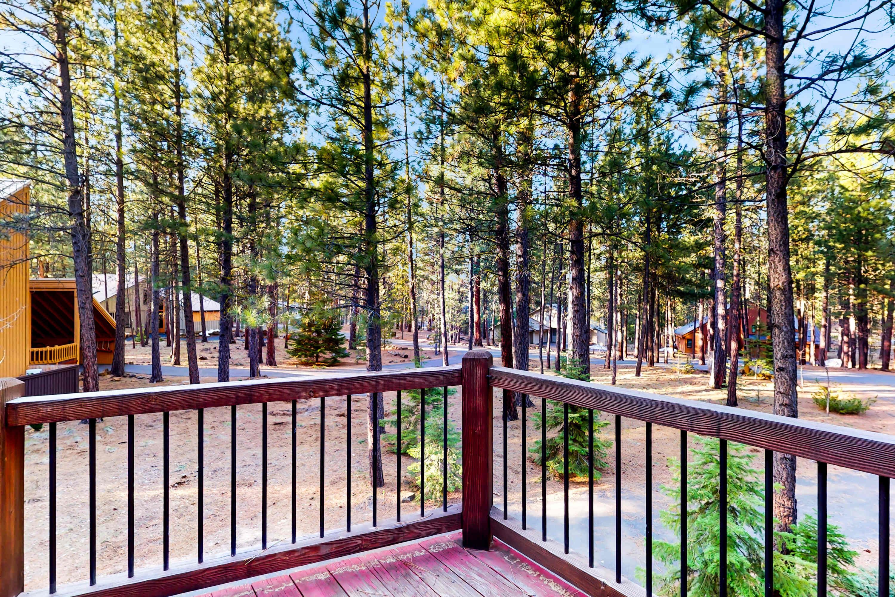 A wooden deck with a metal railing overlooking a forested area with tall trees and a building in the background.