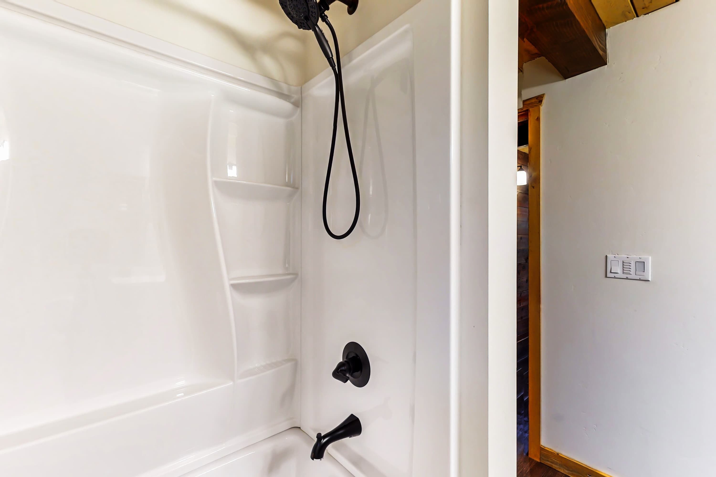 A white bathtub with a showerhead, a small shelf, and a wall-mounted light switch.