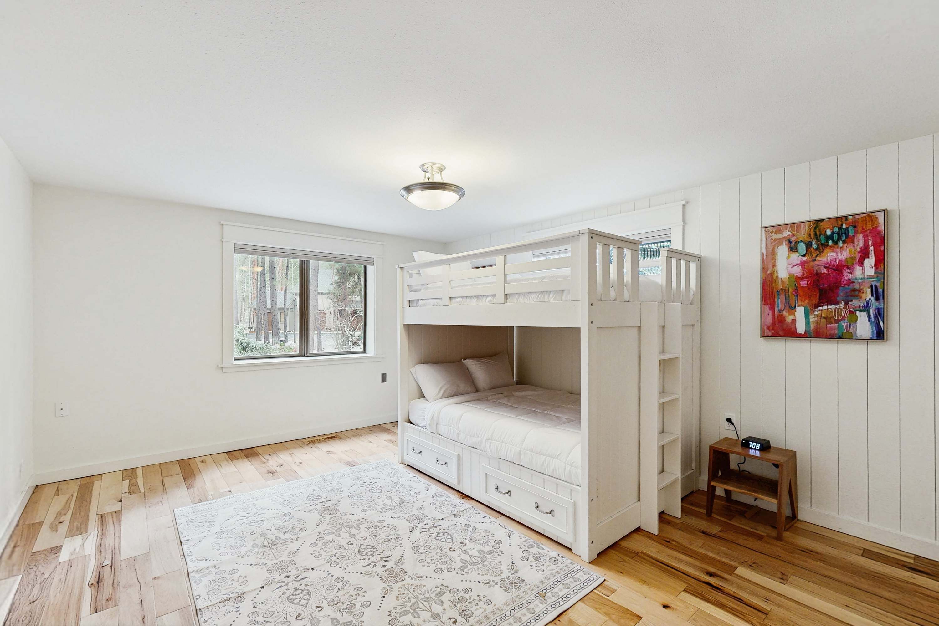 A room with a bunk bed, a window, a small table with a clock, and a colorful painting on the wall.