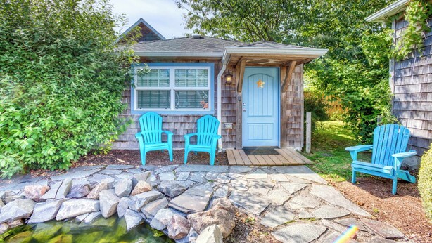 Una piccola casa di legno con una porta blu, due sedie Adirondack blu e un sentiero di pietra che conduce a uno stagno.
