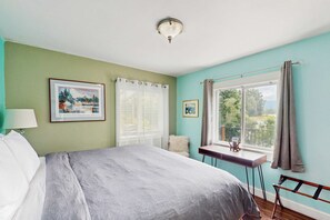 A bedroom with a bed, a window with curtains, a desk with a lamp, and framed pictures on the wall.
