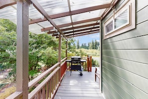 A covered deck with a grill, wooden railing, and a view of a green house.
