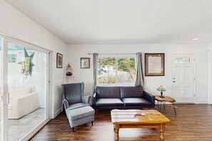 A living room with a sofa, armchair, and coffee table, large windows with curtains, and a door.