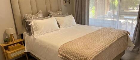 A spacious bedroom with a large bed, a bedside table with a lamp, and a view of the outdoors through a large window.
