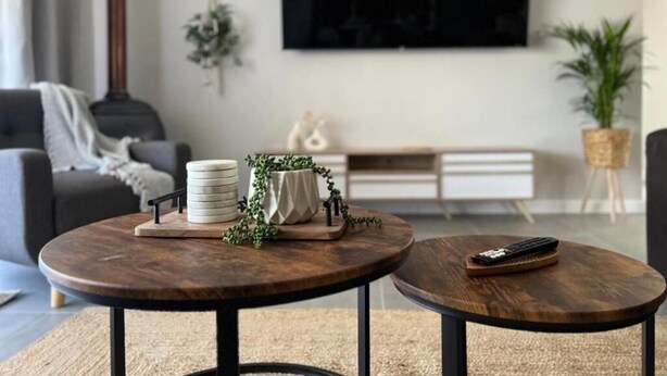 A living room with two round wooden tables, a grey sofa, a TV mounted on the wall, and a potted plant. - Rissie's 3 Bedroom Apartment (Cape Town)