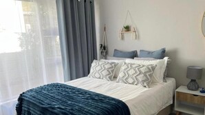 A bedroom with a bed, a bedside table, a hanging macrame plant, and a view of the outside. - Rissie's 3 Bedroom Apartment (Cape Town)