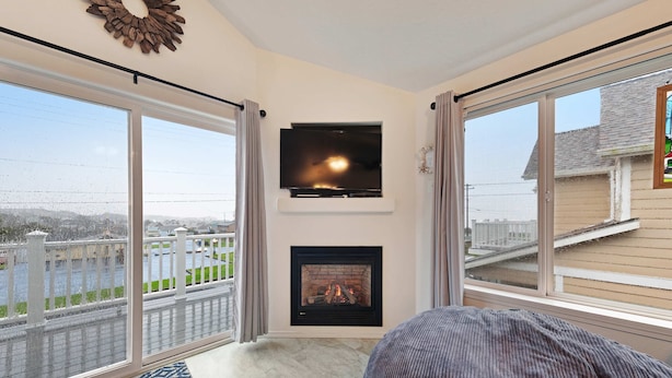 A room with a fireplace, a television, and large windows overlooking a balcony. - Modern Beach House with Partial Ocean Views - Walk to Nye Beach & Attractions (Newport)